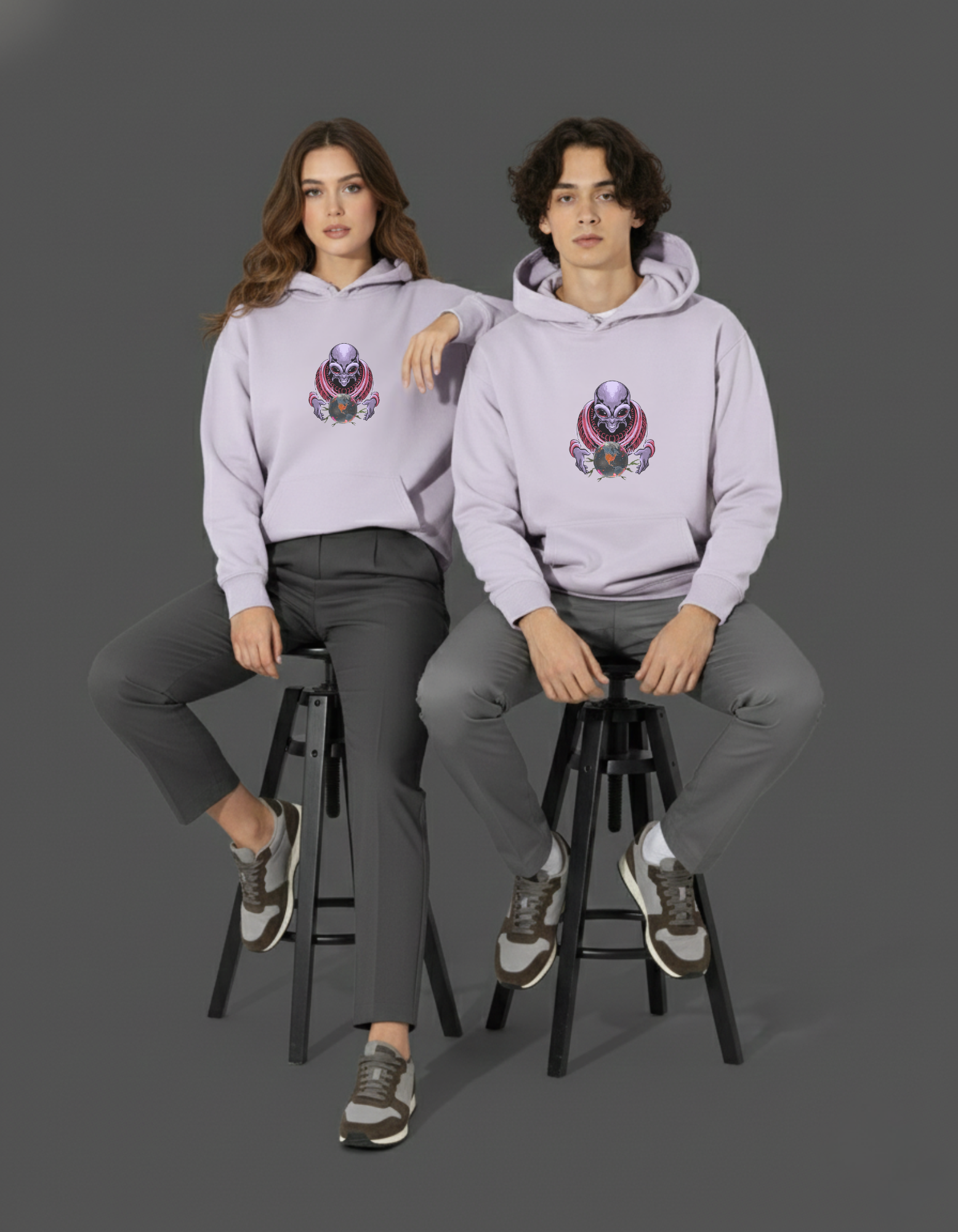 Two people wearing light purple hoodies with a alien with crystal graphic, sitting on stools against a dark background.