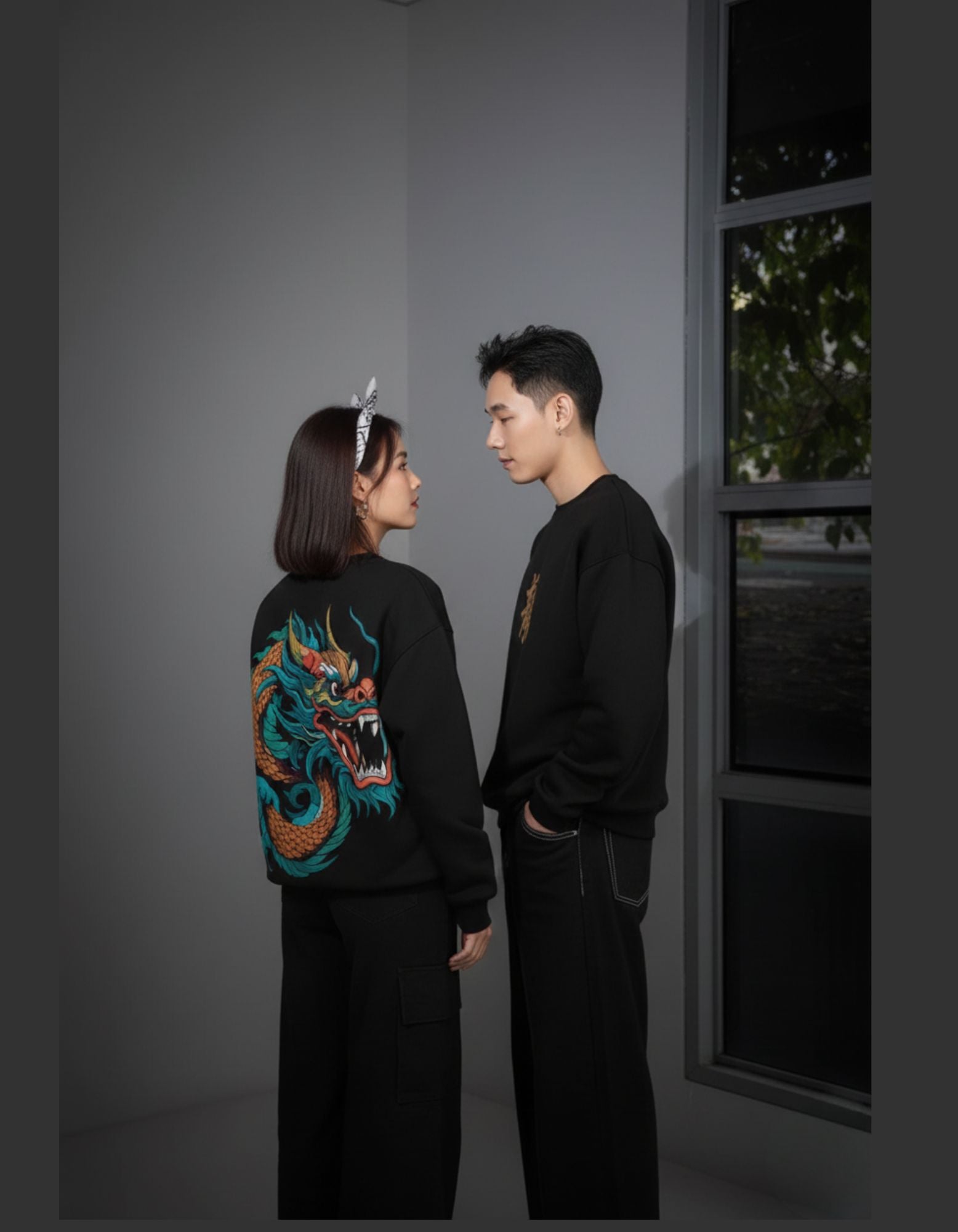 Two people, one with a dragon design on a black shirt, standing against a gray wall.