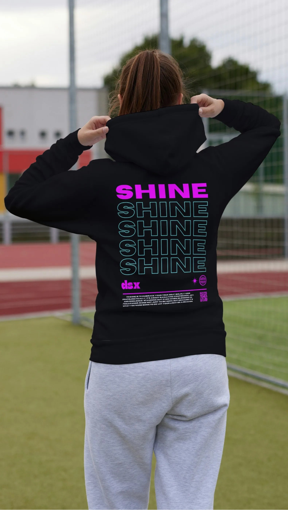 Person wearing a black hoodie with 'SHINE' text on the back, standing on a sports field.